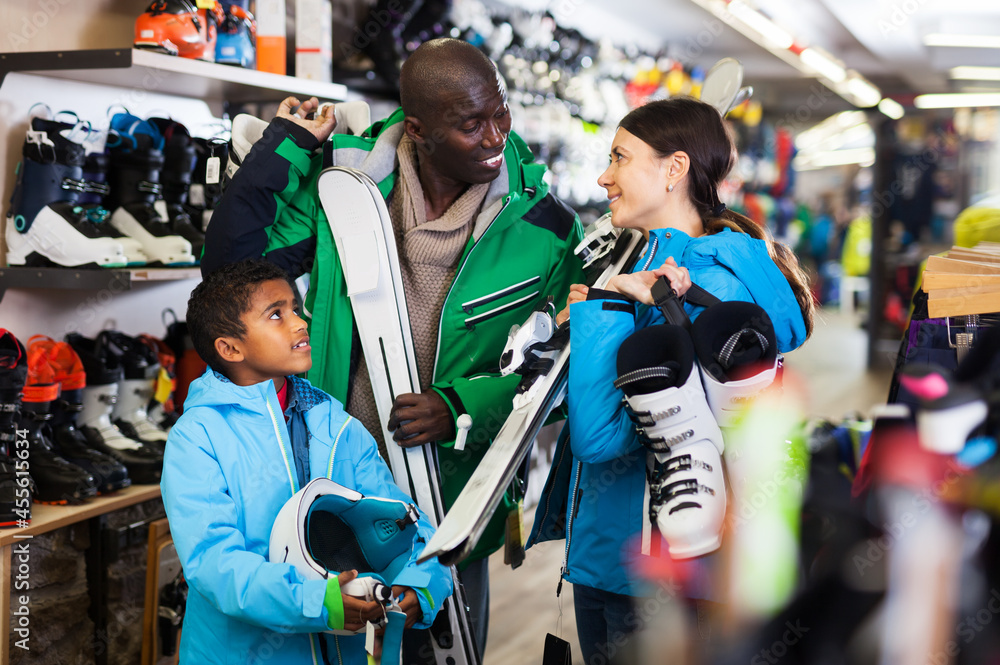 Positive young African man and European woman with preteen son choosing together ski boots for skiing in store of sports gear