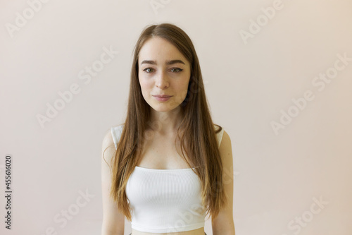 Horizontal studio shot of young beautiful Caucasian woman with dark-blonde straight hair, eye lined, wearing white wide straped crop top, posing against light wall with positive face expression