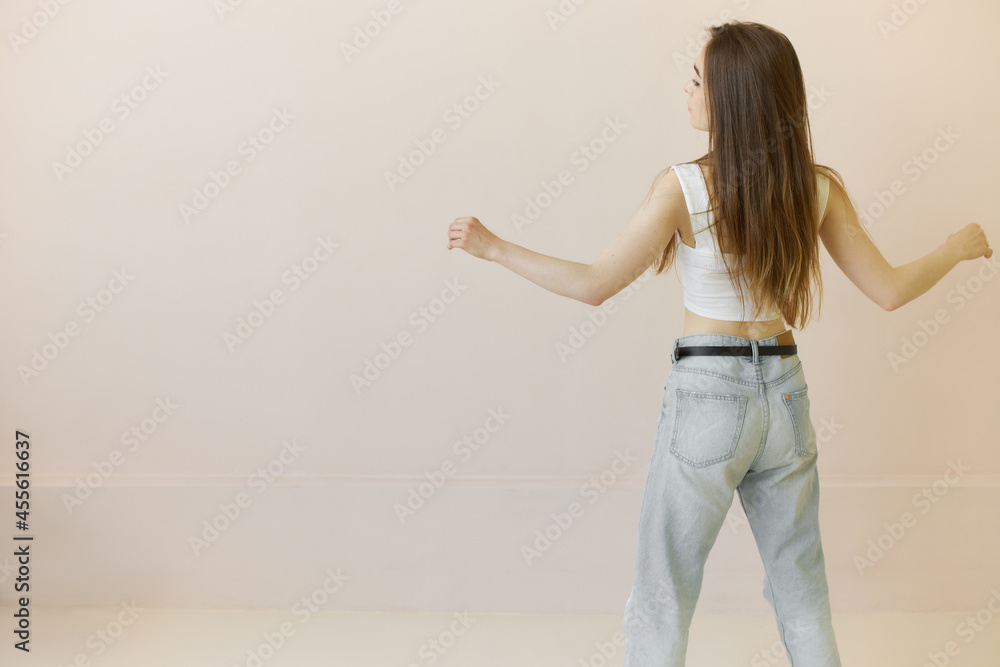 Rare view of Caucasian girl with long hair turned back to camera with hands outstretched to side, moving in dance, during rehearsal, dressed in blue baggy jeans with thin black belt and white tank top
