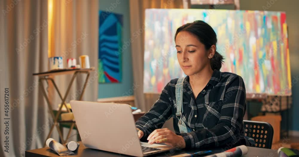 A smiling brunette sits at a desk in front of a laptop in art studio and displays paintings for sale, around her lies a brush, watercolor paints in a tube, in the background a multi-colored painting A smiling brunette sits at a desk in front of a laptop in art studio and displays paintings for sale, around her lies a brush, watercolor paints in a tube, in the background a multi-colored painting