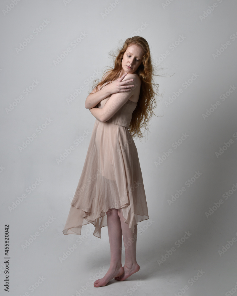 Full length portrait of pretty red haired woman dancer,  wearing skin toned flowing fairy dress. Standing gestural poses  isolated on studio background.