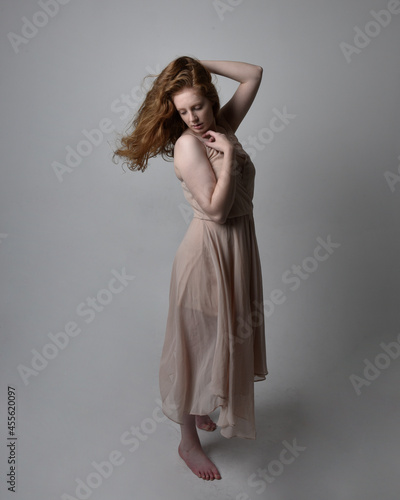 Full length portrait of pretty red haired woman dancer, wearing skin toned flowing fairy dress. Standing gestural poses isolated on studio background.