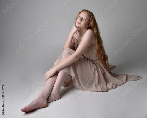 Full length portrait of pretty red haired woman dancer,  wearing skin toned flowing fairy dress. Sitting gestural poses  isolated on studio background.