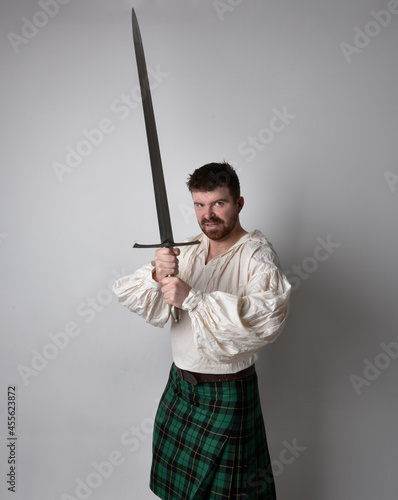 Close up portrait of handsome brunette man wearing Scottish kilt and renaissance white pirate blouse shirt. Holding a sword weapon, action pose isolated against studio background. 
