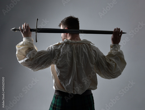 Close up portrait of handsome brunette man wearing Scottish kilt and renaissance white pirate blouse shirt. Holding a sword weapon, action pose isolated against studio background. 