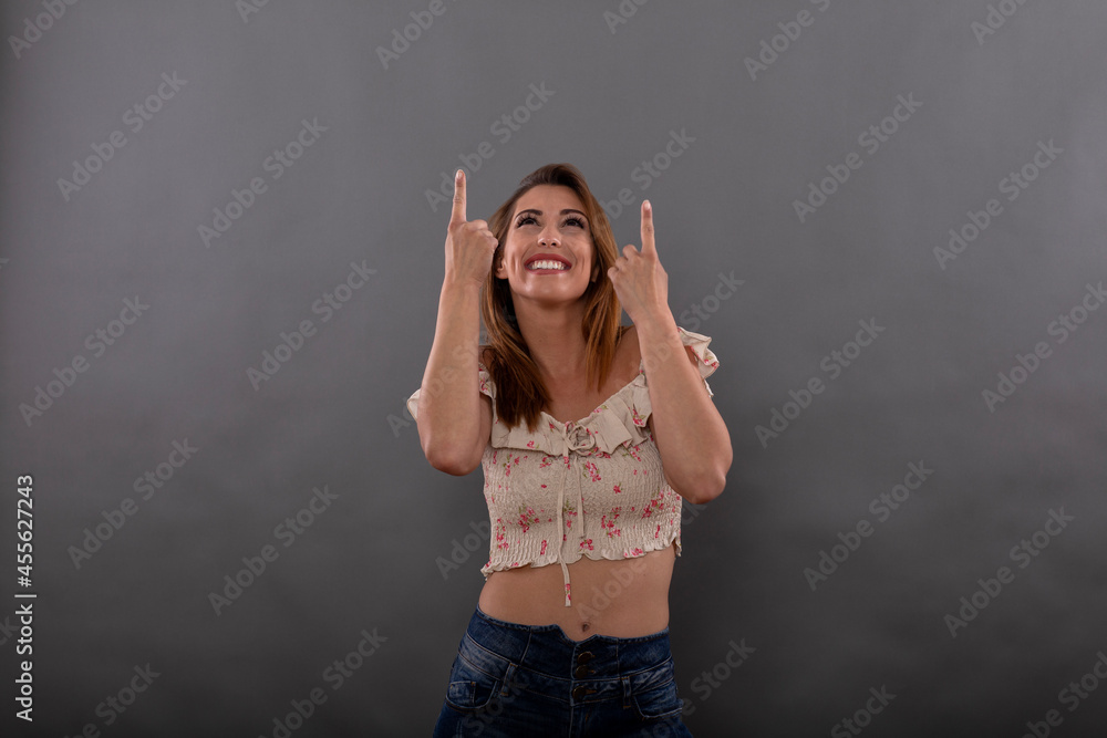 Cheerful brunette model dressed in casual clothes smiles and points upwards with finger isolated on gray background Cheerful brunette model dressed in casual clothes smiles and points upwards with finger isolated on gray background