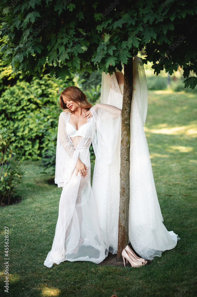 Pretty bride posing near wedding dress that hanging on tree