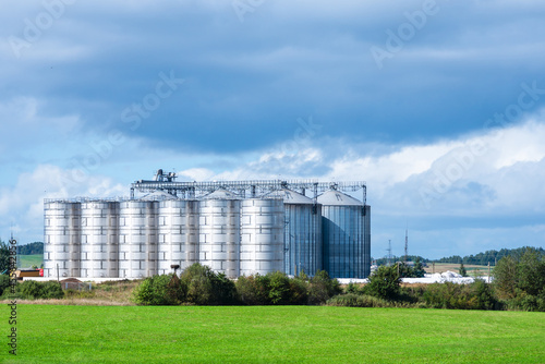 Granary large modern agro-processing plant storage and processing of grain crops. Large metal barrels of grain.Summer cloudy day green field landscape.Copy space.