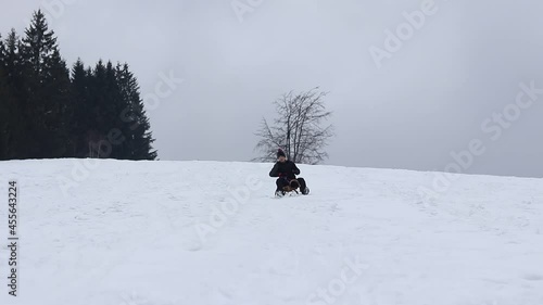 Man from the village in black winter clothes rides down the slope on a wooden sledge. He's trying to correct the direction with his feet.  Winter fun. 23-year-old boy. Going back to his childhood photo