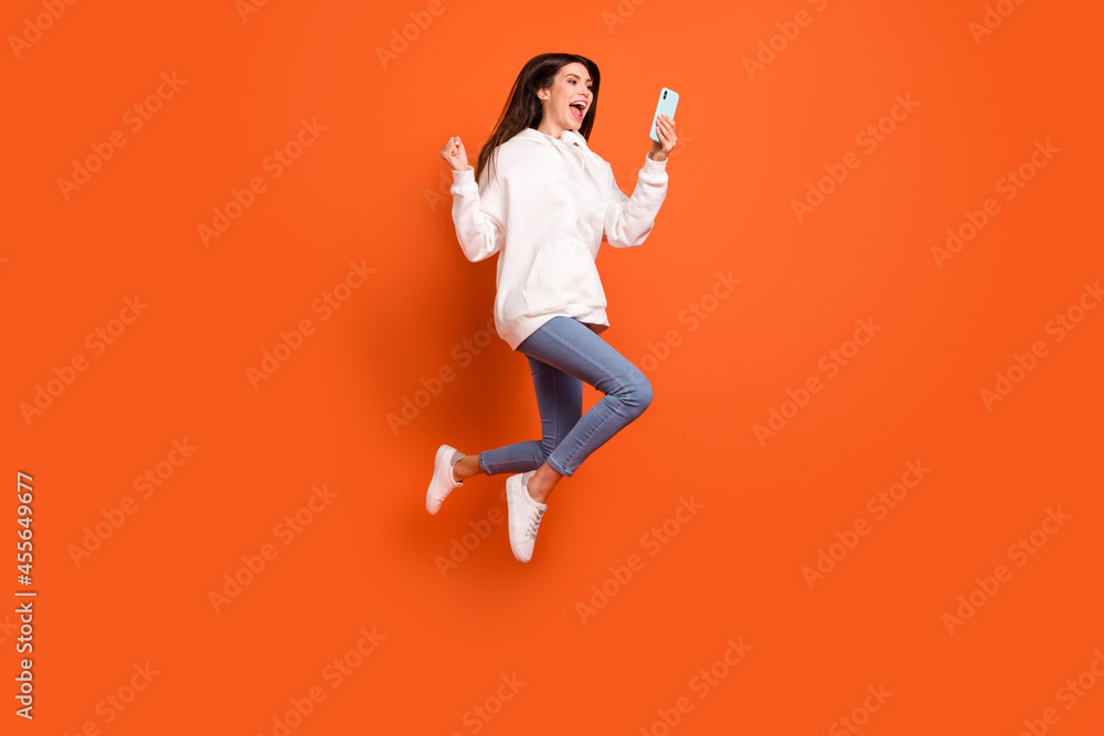 Full length body size view of pretty trendy lucky cheerful girl jumping using device win isolated over bright orange color background Full length body size view of pretty trendy lucky cheerful girl jumping using device win isolated over bright orange color background