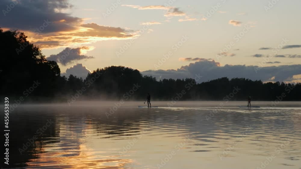 Evening, the silhouette of a man and boy on stand-up paddleboards. Paddling on the quiet lake after sunset. Evaporating water.