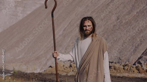 Jesus Christ with a staff in robe stands on the mountain and looks into the distance. There are bruises on his face, wounds on his hands.