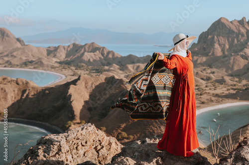 Happy Asian woman in summer hat and orange dress standing on top hill and holding Indonesia traditional fabric called kain songket with background of hills and seascape at Padar Island Labuan Bajo