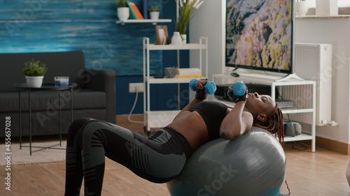 Fototapeta Naklejka Na Ścianę i Meble -  Slim woman with black skin exercising body muscle doing morning exercices with dumbbells sitting on fitness swiss ball during yoga workout in living room. Fit athlete adult wearing sportswear