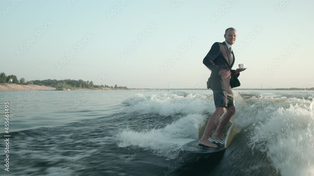 A surfer jumping on a wakeboard in a suit with a cup of tea or coffee in his hands. An experienced wakeboarder sprays water drops into the camera. A surfer jumping on a wakeboard in a suit with a cup of tea or coffee in his hands. An experienced wakeboarder sprays water drops into the camera.