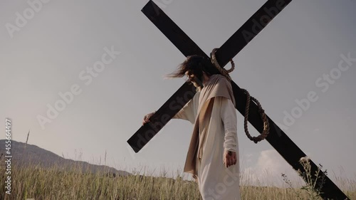 Jesus carries the cross on his back. He walks on the grass, against the background of the hills. The wind is blowing, the sun is shining.