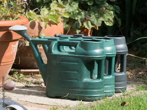  Three green watering cans stand side by side on slabs ready for watering a garden.Flower pots behind and grass in foreground