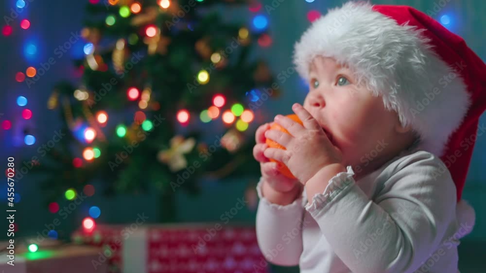 Christmas Eve. Portrait of caucasian toddler girl in xmas hat eating tangerine. Side view. Slow motion. The concept of winter seasonal holidays. Christmas Eve. Portrait of caucasian toddler girl in xmas hat eating tangerine. Side view. Slow motion. The concept of winter seasonal holidays.