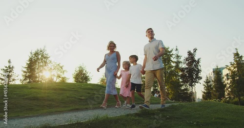 A friendly family strolls along the path in the park in the evening. They walk hand in hand. Mother and father are at the edges, and in the center are children. Around them are green grass and trees