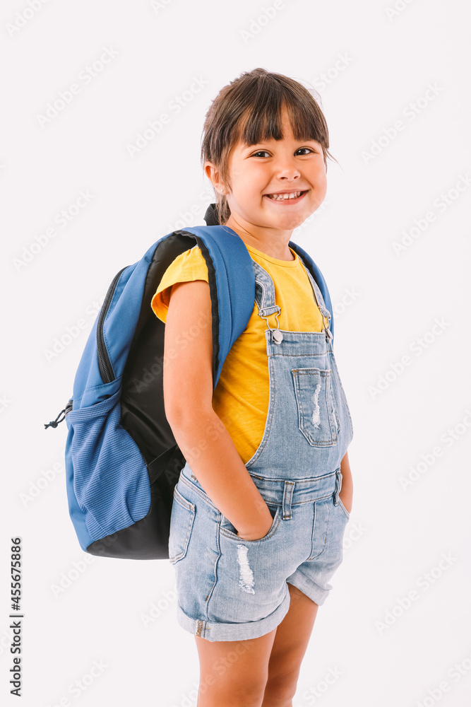 Little girl with black hair dressed in a denim overalls and a blue t-shirt, with a backpack ready for going back to school, on her side, on white background Little girl with black hair dressed in a denim overalls and a blue t-shirt, with a backpack ready for going back to school, on her side, on white background