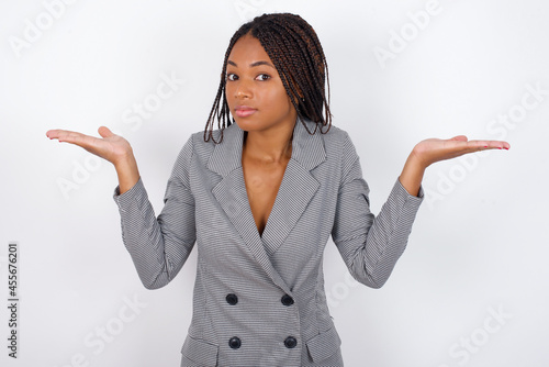 Obraz na płótnie Puzzled and clueless Young african american business woman with braids over white wall with arms out, shrugging shoulders, saying: who cares, so what, I don't know