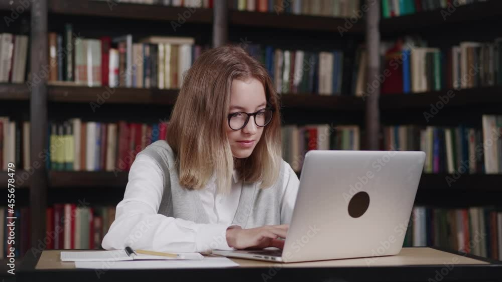 Girl with short hair wearing glasses working in the school library. She uses the computer. Girl with short hair wearing glasses working in the school library. She uses the computer.
