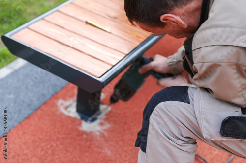 Blur image of installation of benches on the playground. Bench and holes on artificial grass for fitting, mounting. A man in overalls drills a hole with a drill for a screw.