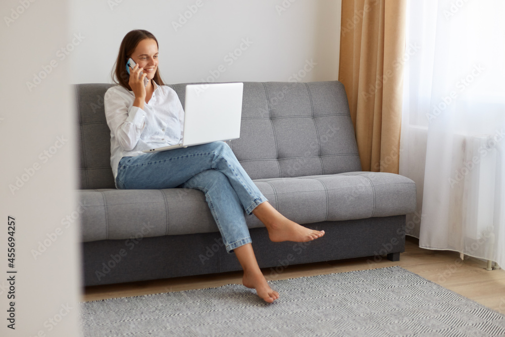 Portrait of beautiful dark haired woman speaking on cellphone in front of her laptop computer at home while sitting on sofa in light living room, having positive facial expression. Portrait of beautiful dark haired woman speaking on cellphone in front of her laptop computer at home while sitting on sofa in light living room, having positive facial expression.