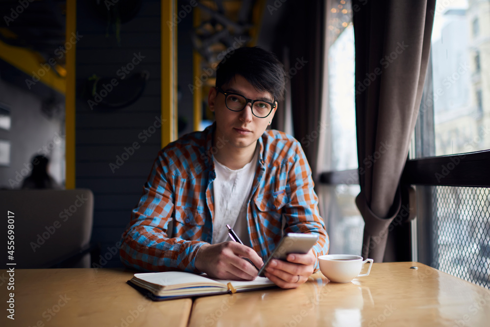 Millennial mobile phone user sitting at the table with notebook while preparing for exams. Smartphone application, modern technology for learning Millennial mobile phone user sitting at the table with notebook while preparing for exams. Smartphone application, modern technology for learning