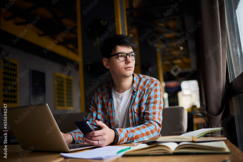 Fototapeta premium Millennial male using mobile phone while sitting at the table with books while front open laptop computer. Smartphone application. Working on freelance, using technology
