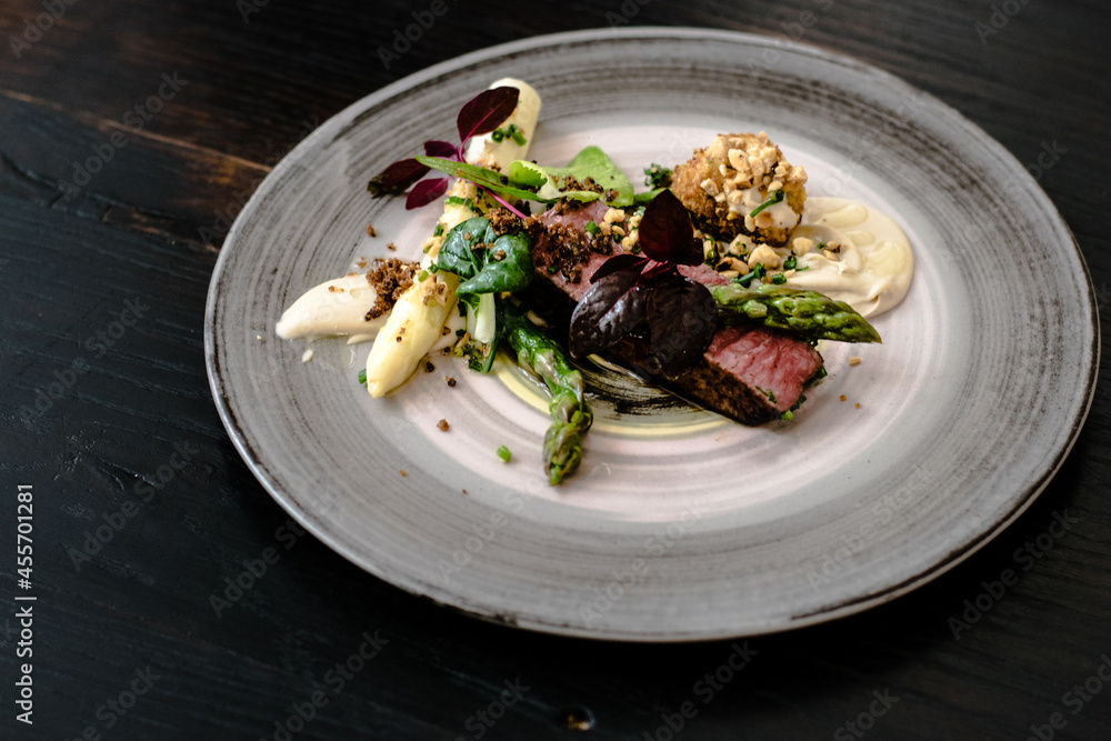 Dish of ostrich steak with white and green asparagus, garden greens and sauce on a plate on wooden table in a restaurant Dish of ostrich steak with white and green asparagus, garden greens and sauce on a plate on wooden table in a restaurant