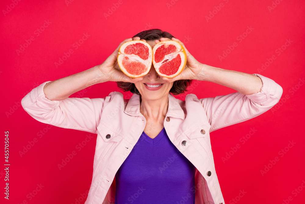 Photo of pensioner lady hold two grapefruit slice cover eyes wear pink jacket isolated red color background