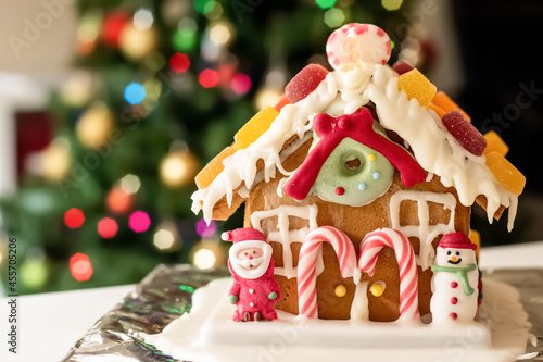 Homemade gingerbread house decorated with icing, sweets , figurines, lollies and candy canes with blurred Christmas tree on background