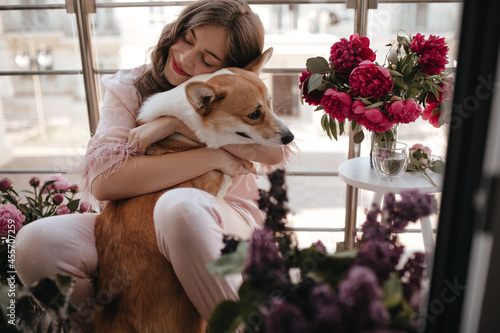 Young ukrainian girl sincerely hugging the dog in the peonies garden. Bunette lady with natural make up and closed eyes sitting on the floor of the french balcony in light pink stylish clothes photo