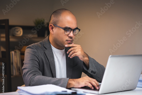 Young asian businessman working with laptop computer in the dark office at night. Attractive Indian man work hard overtime serious thinking in home with floor lamp ambient warm light late at night