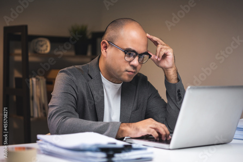 Young asian businessman working with laptop computer in the dark office at night. Attractive Indian man work hard overtime serious thinking in home with floor lamp ambient warm light late at night © Chanakon