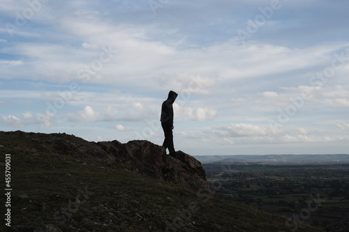 A hooded figure walking towards the top of a rocky outcrop. Looking down across the landscape