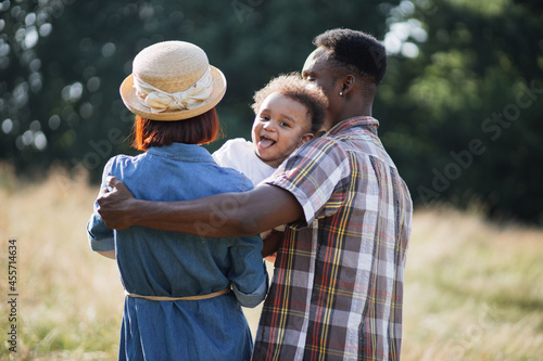 Φωτογραφία Back view of multiracial parents standing in hugs on summer field and holding cute son on hands that showing tongue on camera