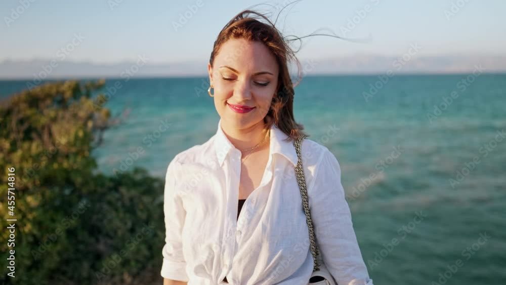 Portrait of smiling woman, she standing on cliff beach, enjoying wind, fresh air. Blue sea background. Lady in white linen shirt. Travel destination, vacation in Greece. Portrait of smiling woman, she standing on cliff beach, enjoying wind, fresh air. Blue sea background. Lady in white linen shirt. Travel destination, vacation in Greece.