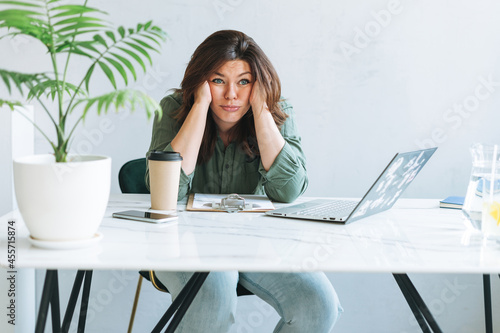Young thinking unhappy brunette woman plus size working at laptop on table with house plant in the bright modern office