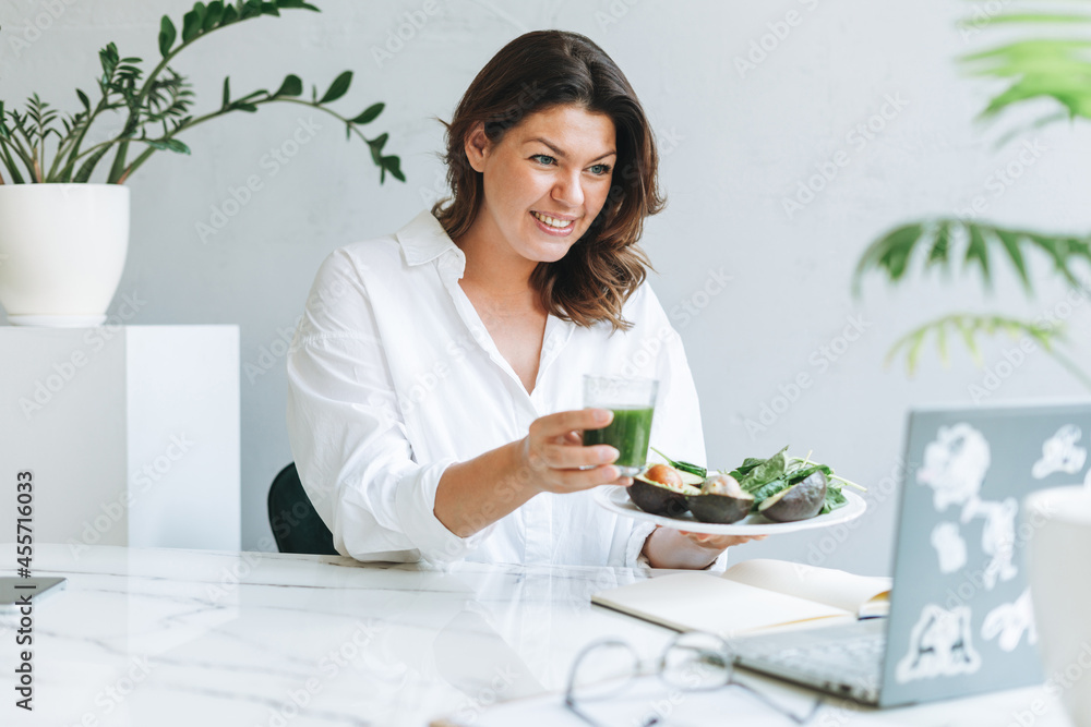 Young smiling brunette woman nutritionist doctor plus size in white shirt with green healthy food at modern bright office room. Doctor communicates with patient online