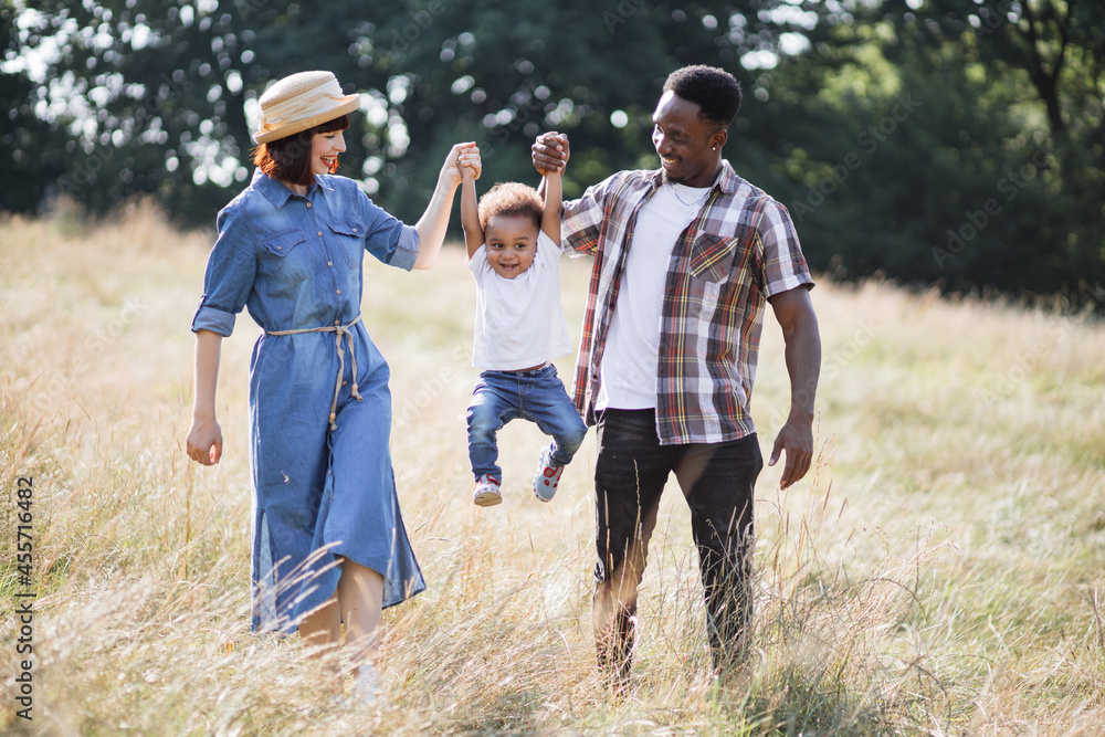 © sofiko14 - Cheerful young parents raising son up for both hands while playing together on summer field. Multiracial family in casual outfit enjoying every moment spending together.
