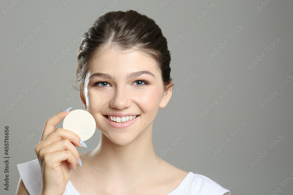 Beautiful girl with foundation smear on her face holding sponge against grey background Beautiful girl with foundation smear on her face holding sponge against grey background
