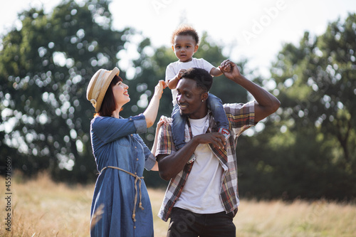 Beautiful multicultural family of three dressed in casual clothes walking together of summer field. African man holding boy in shoulders, caucasian woman embracing them.