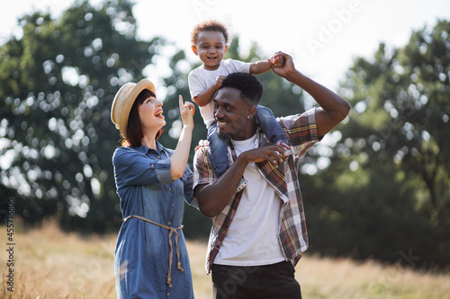 Little african boy sitting on father's shoulders and smiling sincerely to caucasian mother that walking near. Happy multiracial family outdoors. Summer time concept.