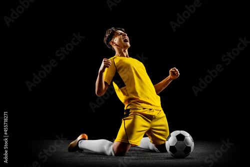 Full-length horizontal portrait of young man, male soccer football player training isolated on black background. photo