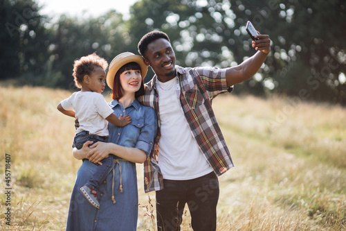 Pretty caucasian mother holding cute son on hands while african american father taking selfie on modern smartphone. Concept of family, technology and outdoors activity.