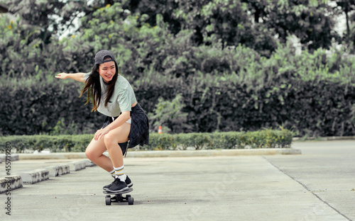Portrait beautiful sportive Asian female skater wearing hipster shirt with shorts, smiling with happiness, standing on skateboard and playing outdoor with copy space. Activity and Adventure Concept.