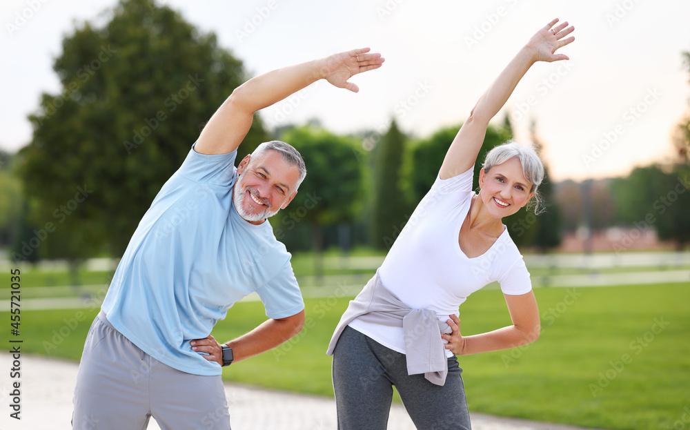 Happy smiling mature man and woman in sportswear stretching body while warming up together outdoors Happy smiling mature man and woman in sportswear stretching body while warming up together outdoors