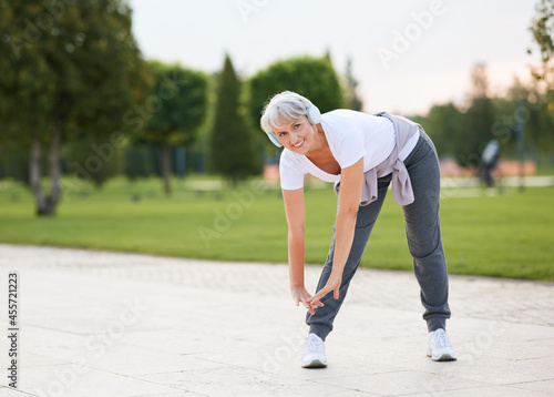 Elderly fit woman in wireless headphones doing stretching exercises before jogging outside